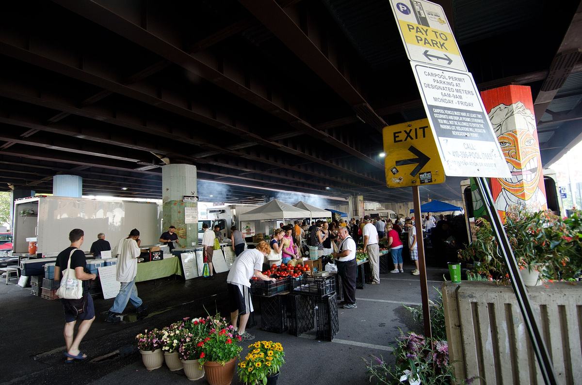 Shoppers browsing produce crates and potted flowers at an outdoor market held in a parking lot beneath a highway interchange