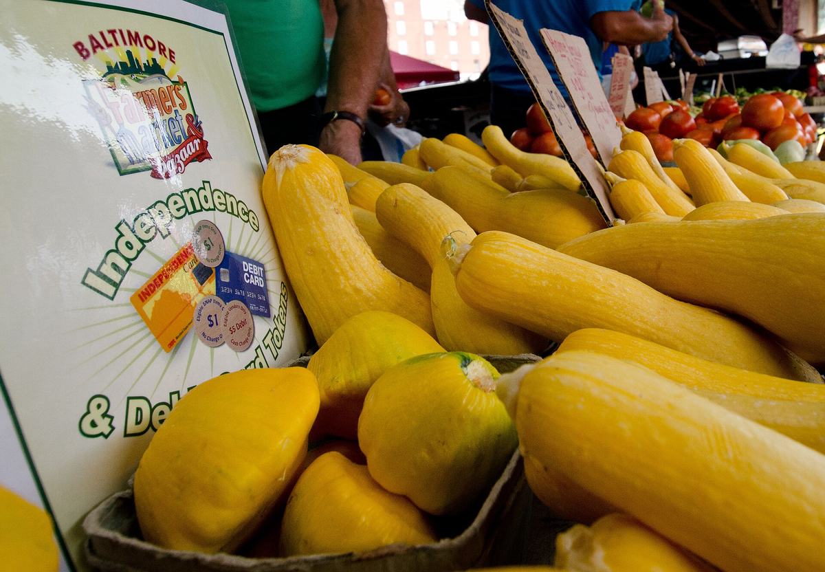 A market stall sign showing an EBT Independence Card alongside $1 and $5 wooden tokens, beside crates of yellow squash and tomatoes